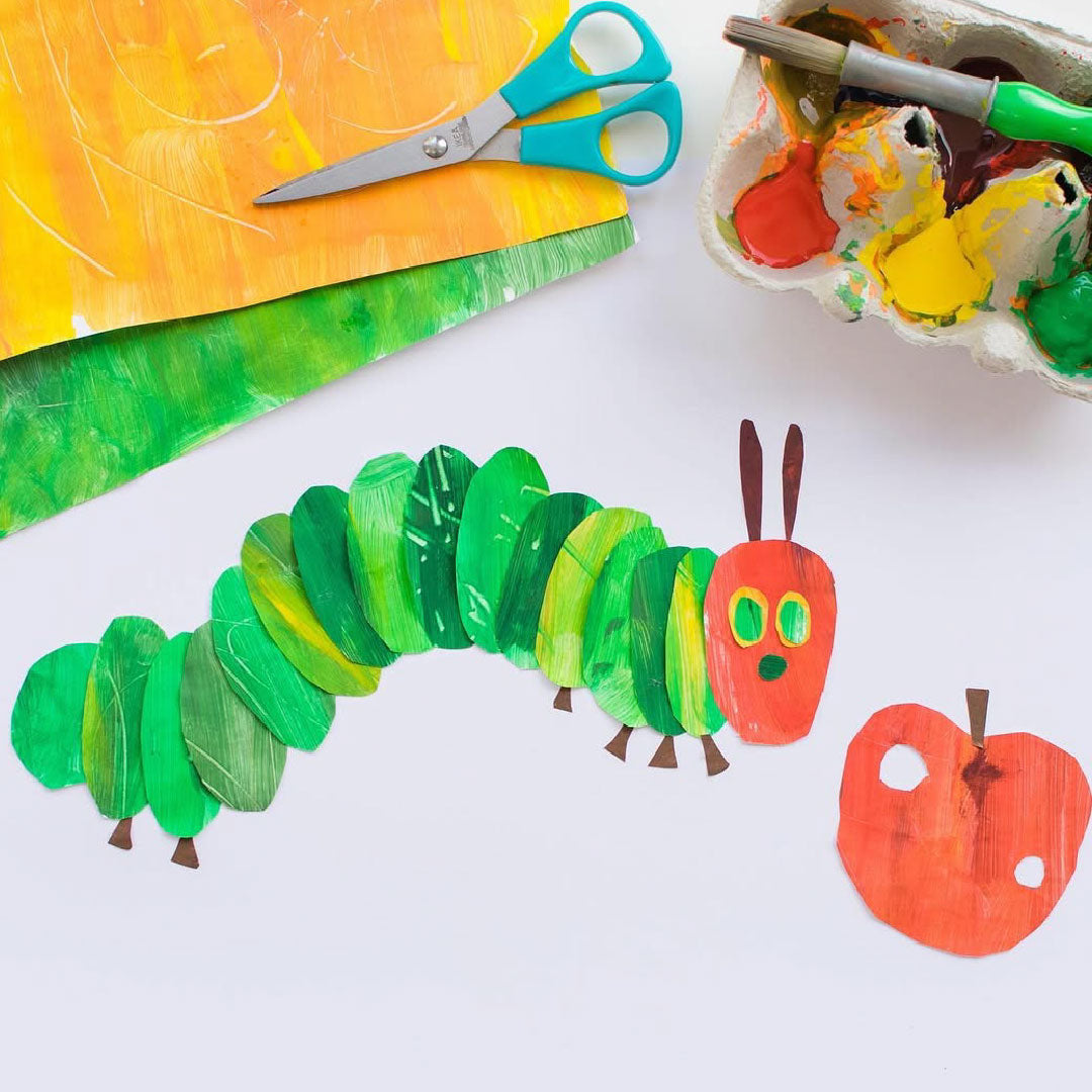 A painted paper craft of a green caterpillar and a red apple surrounded by scissors, painted paper sheets, and an egg-carton paint palette.