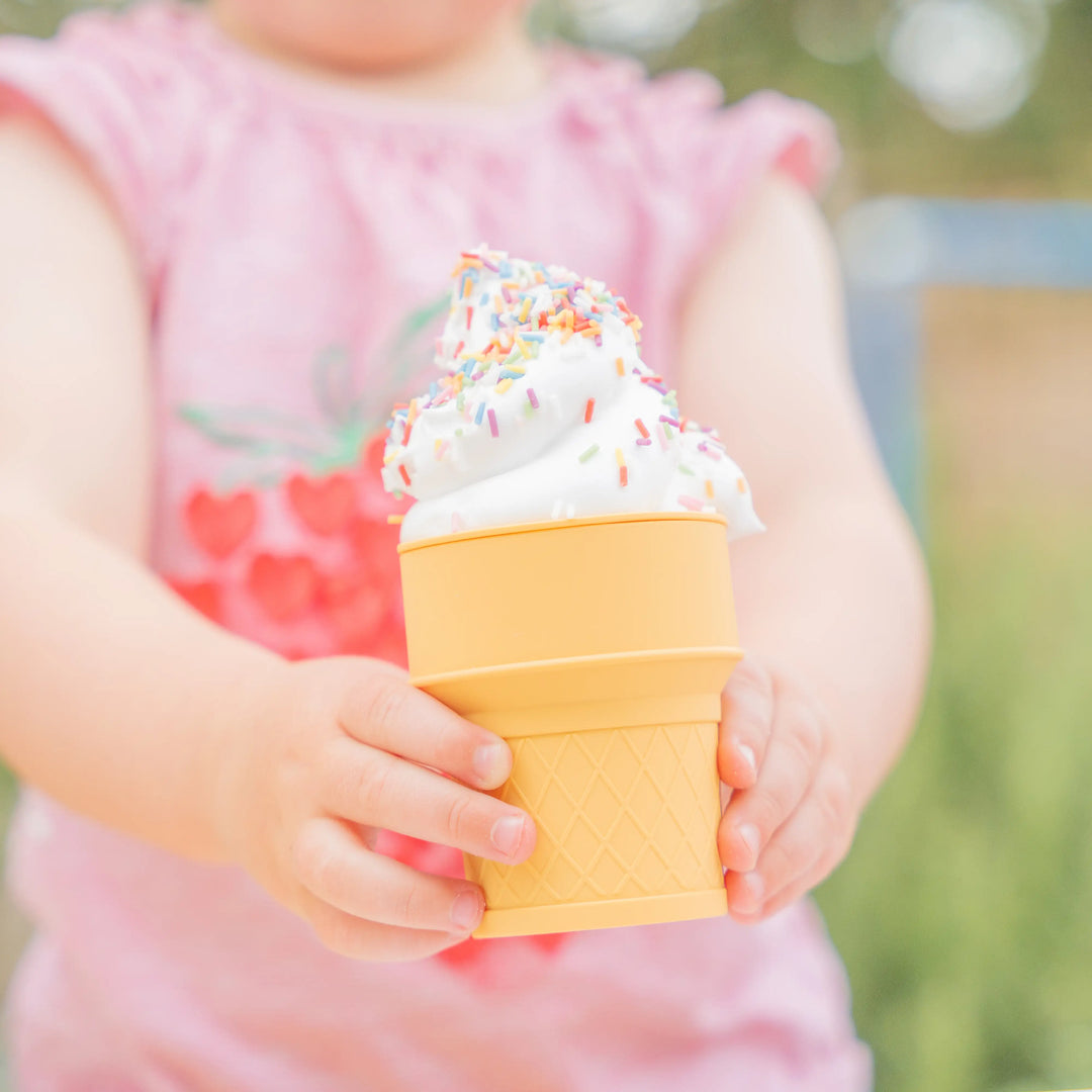 A child holding a beige silicone toy ice cream cone filled with shaving foam and topped with sprinkles.