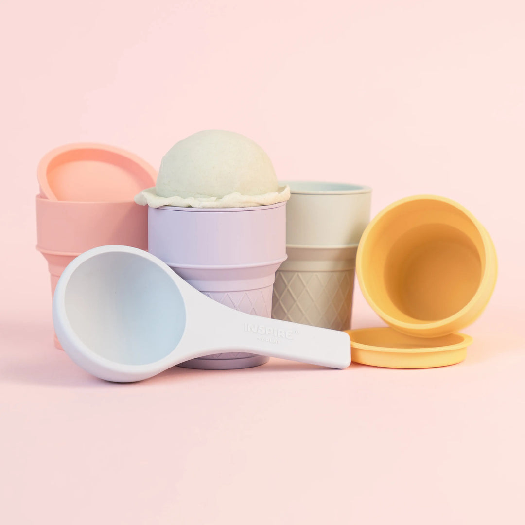 A set of pastel-coloured ice cream cones with matching coloured lids and a blue silicone scoop on a soft pink background. The set includes toy ice cream cones in pink, lavender, green and yellow, along with a blue scoop.