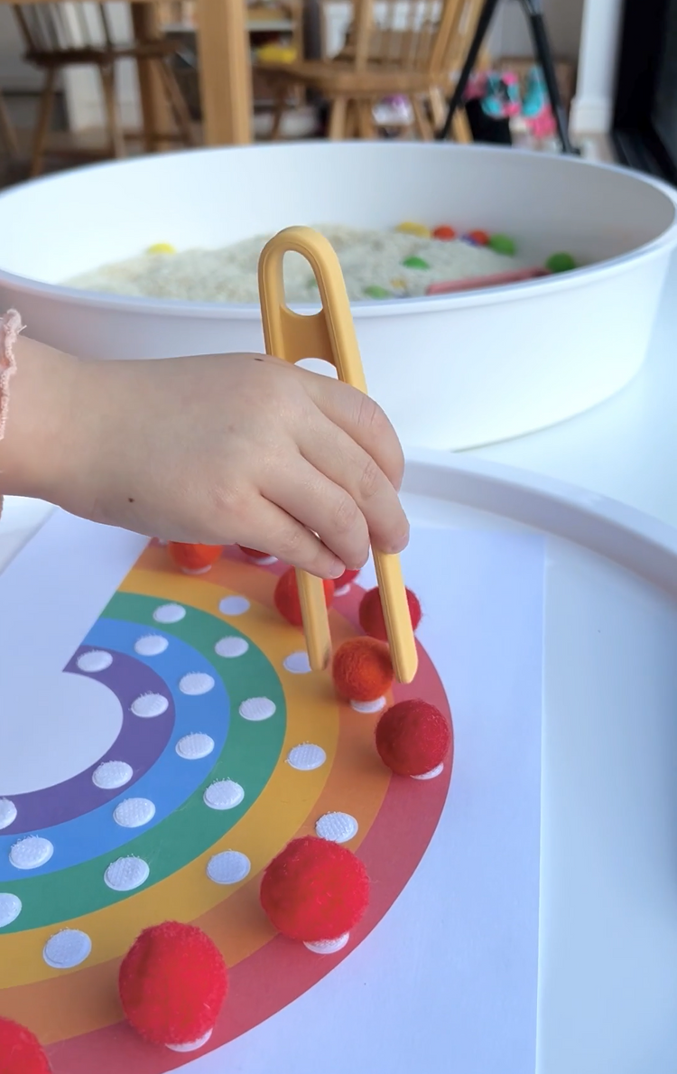 Child retrieving pompoms from a PlayTRAY filled with rice and placing them onto a rainbow activity sheet using sensory play tongs.