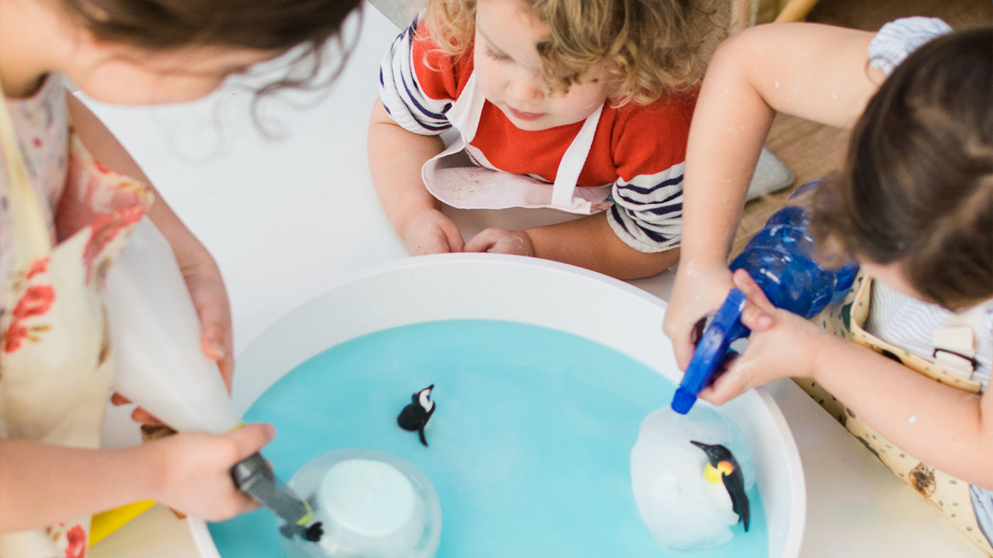 3 young children playing in a play tray with ice and penguin figurines